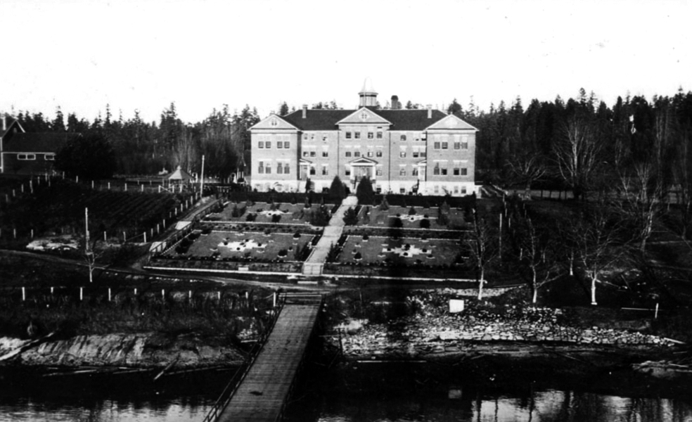Aerial black and white photograph overlooks a wooden dock leading from the shoreline to a stepped walkway through manicured symmetrical gardens fronting a massive, imposing three-story brick building.