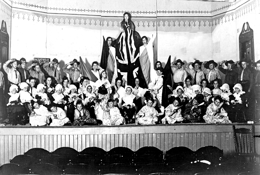 Black and white photograph of a large group of child actors in costume arranged on a stage in three rows, kneeling at the front, sitting in the middle and standing at back. The back row features in its centre three actresses elevated from the rest, each holding flags vertically with a central Union Jack above all.