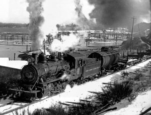 Black and white photograph of a steam engine, smoke and steam billowing from its chimney and steam from its whistle, pulling a line of empty flat cars. The waters of Ladysmith Harbour, with pile-driven stakes demarking booming grounds, form the backdrop.