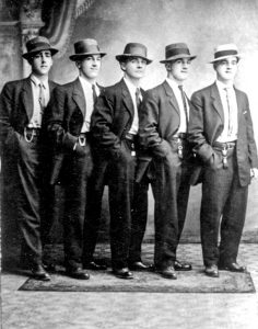 Black and white photograph of five young men smartly dressed in two piece suits, white shirt and tie with period hats, standing en echelon at an angle to the camera, right hands in their pants pockets.