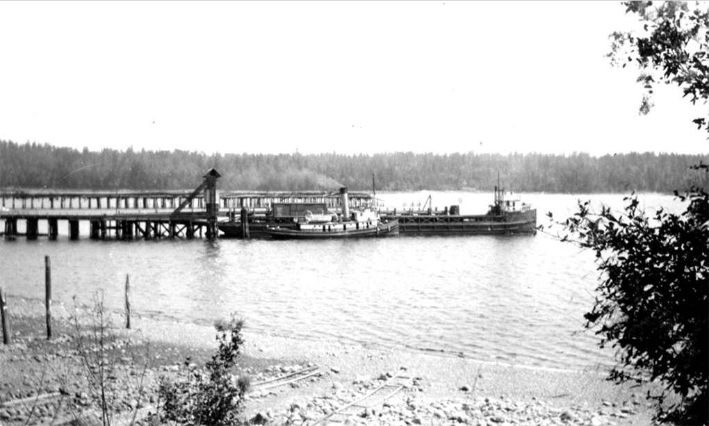 Black and white photograph of a single funnelled steam tug nosing a partially loaded barge alongside a wharf.