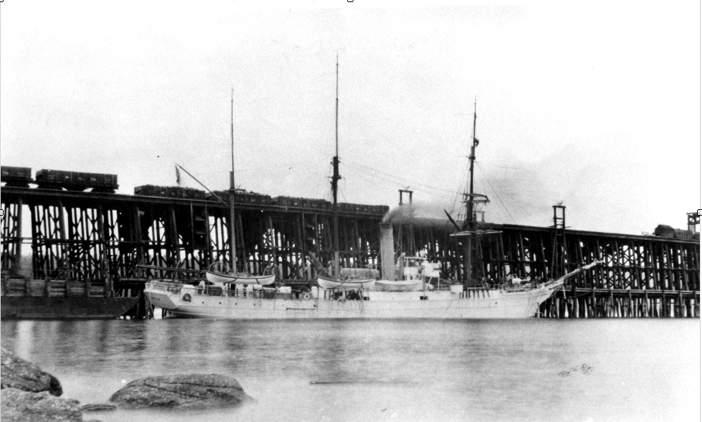 Black and white photograph of a three masted ship, sails furled, with prominent smoking funnel amidships moored beside and dwarfed by pilings of a large wooden wharf, atop of which are loaded and empty wooden coal railcars.