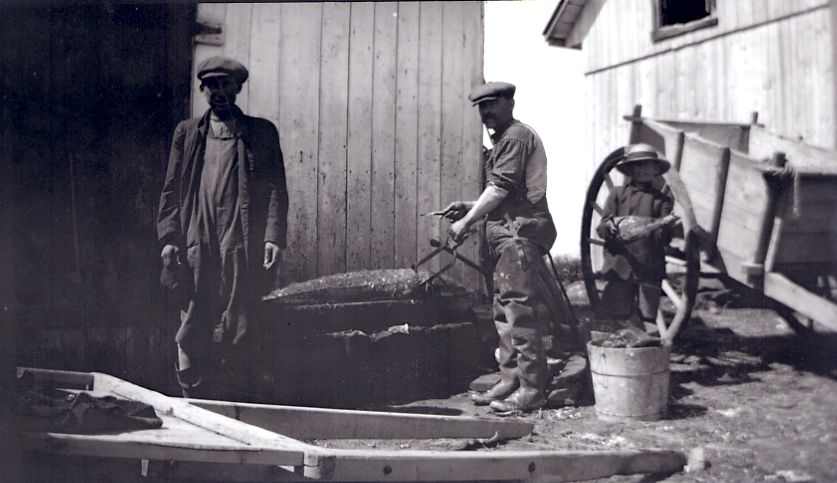 Black and white photograph of two men flensing a sturgeon. Centre: man with a tool working on the long fish. Left: man looking at the camera. Right: child near a wooden cart. Outdoors, building and metal bucket in the foreground on the right.