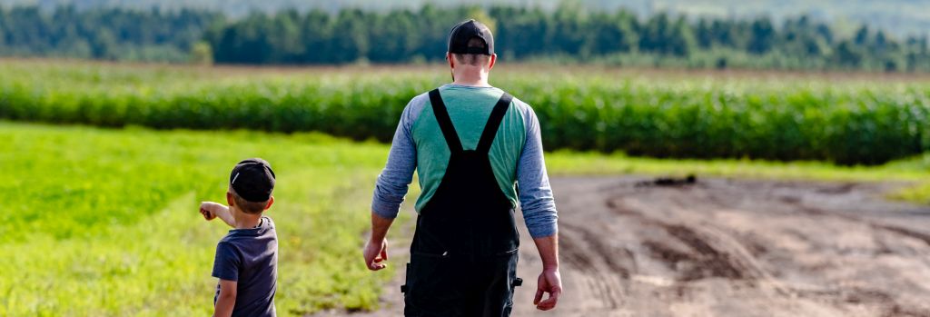 Rear view of an adult and a child walking on a dirt path through a lush green field. The child is pointing to the left.