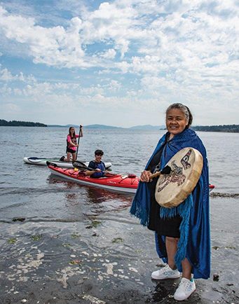 Colour photograph of a Coast Salish woman with long salt and pepper colored hair swept back off her face wearing an azure colored cape closed at the neck over a black knee length dress and white sneakers. She holds in her right hand a drum stick and in her left a round skin drum decorated with Coast Salish stylised animals painted in black and red. She stands on a rocky shore. Behind and to the left of her a person sits in a red and white kayak, and another kneeling on a light colored paddle board. Forested islands form the skyline.