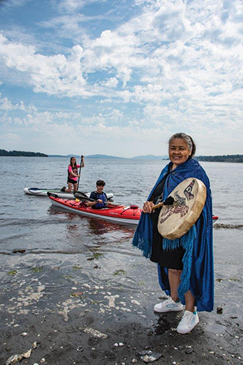 Colour photograph of a Coast Salish woman with long salt and pepper colored hair swept back off her face wearing an azure colored cape closed at the neck over a black knee length dress and white sneakers. She holds in her right hand a drum stick and in her left a round skin drum decorated with Coast Salish stylised animals painted in black and red. She stands on a rocky shore. Behind and to the left of her a person sits in a red and white kayak, and another kneeling on a light colored paddle board. Forested islands form the skyline.