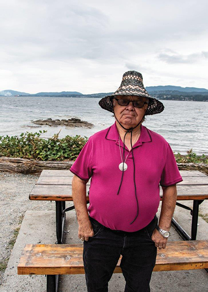 Colour photograph of a man wearing a First Nations traditional cedar hat. The ocean and a picnic table is visible behind him.