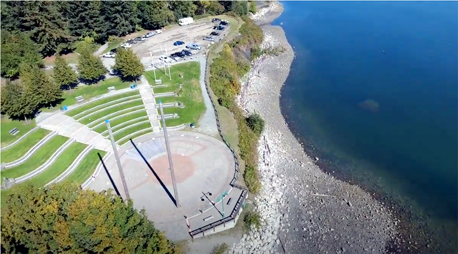 Aerial colour photograph of a circular cement and brick outdoor stage located on a shoreline. Two tall wooden poles stand off-centre on the seaward side of the stage. Landward, facing the stage is a semicircular sloped area sculpted into stepped grass covered benches.