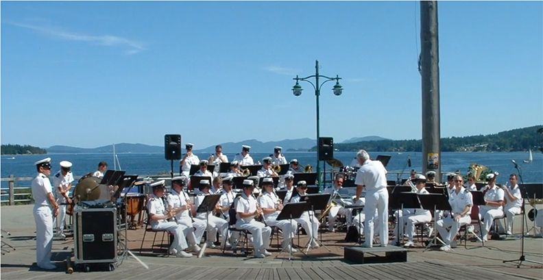 Colour photograph of an approximately 30-member brass band in white uniforms playing on an open air stage, the backdrop to which is a view of sea and wooded islands under a virtually cloudless blue sky.