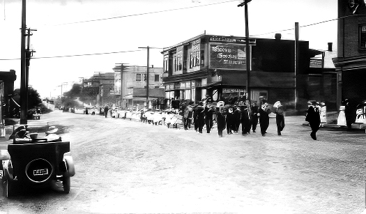 Black and white vintage photograph of an unpaved street lined with two and three story commercial buildings along which a parade is led by a uniformed brass band of about 15 musicians followed by over a hundred children, many dressed in white with white hats, walking in rows three abreast. These followed by a file of vehicles stretching beyond the field of sight.