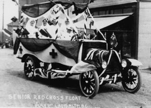 Black and white photograph, oblique view, of a spoke-wheeled open box long wheel based truck bedecked with Union Jack and Canada Red Ensign flags on the hood and running boards. The driver, dressed in white with a white cap, sits at the wheel. Behind him, the box with sides adorned with fabric sporting crosses contains at least five seated nurses in white uniforms including white head scarves.