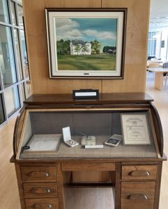 Wooden roll-top desk with a glass display case showing medals, certificates, and documents. Above it hangs a framed painting of a white two-story house with trees in the background. A label reads G.J. Guttormsson Collection.