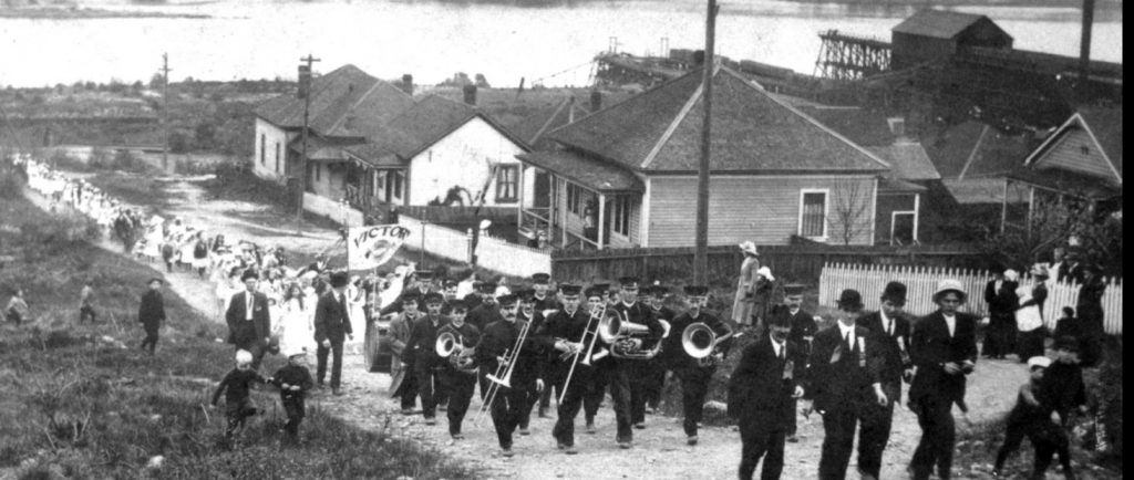 Black and white photograph looking downhill along an unpaved street up which a brass band is leading a procession of women and children. The waters of a harbour form the background.