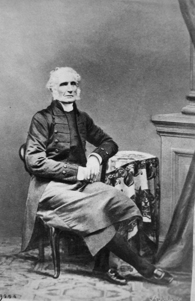 Black and white studio portrait of Bishop George Jehoshaphat Mountain. The elderly man is sitting on a chair wearing the robes of an Anglican priest. The studio decor suggests the interioir of the classic style of the bourgeoisie of the second half of the 19th century.