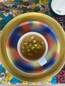 Overhead shot of an orange soup bowl resting on a small plate with blue, red, and yellow patterns, which is itself placed on a large golden plate. The ensemble is on a colorful floral-patterned tablecloth.