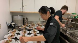 Professional kitchen: two people preparing dishes. Foreground: Asian woman with black hair, black chef's uniform, tongs above identical plates. Background: bearded man with brown hair, black uniform. Atmosphere of concentrated culinary precision.