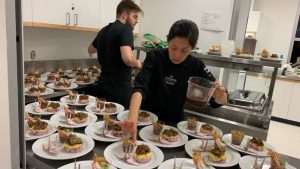 Professional kitchen: two people are plating numerous identical dishes. Foreground: Asian woman with black hair, black chef's uniform, pouring brown liquid onto the plates. Background: bearded man with brown hair, black uniform. Atmosphere of meticulous preparation for an event.