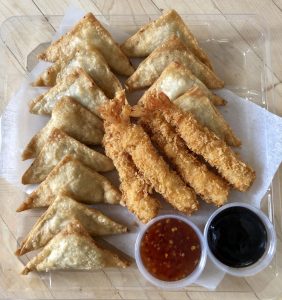 Overhead shot of a dozen triangular fried wontons and four Japanese-style breaded fried shrimp. The items are arranged in a transparent, rectangular plastic container. At the bottom of the dish, there are two small pots of sauce: a red sweet chili sauce and a dark sauce (likely soy or plum). The entire dish rests on a light-colored wooden surface.