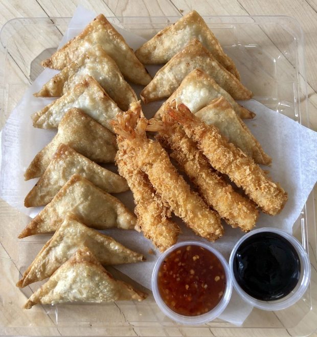 Overhead shot of a dozen triangular fried wontons and four Japanese-style breaded fried shrimp. The items are arranged in a transparent, rectangular plastic container. At the bottom of the dish, there are two small pots of sauce: a red sweet chili sauce and a dark sauce (likely soy or plum). The entire dish rests on a light-colored wooden surface.