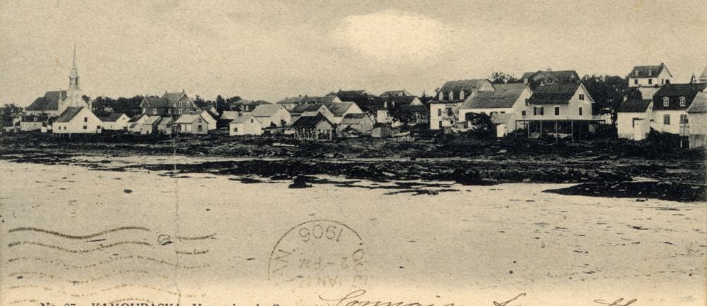 Sepia postcard No. 87. KAMOURASKA. – View from the river (panoramic view of a small coastal town). Foreground: expanse of water (St. Lawrence River). Shore: houses, church with a pointed steeple. Handwritten inscriptions in French. Stamp 1906. Bottom left: Publishers: Pruneau & Kirouac, Quebec.