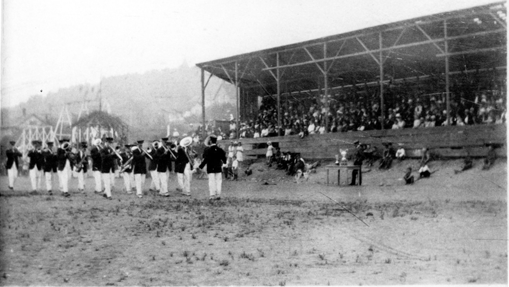 Black and white photograph of members of band playing outside with spectators in grandstand looking on.