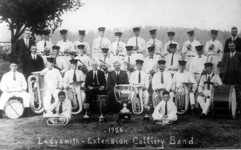 Approximately thirty uniformed band members pose in three rows with their instruments behind four silver trophies.