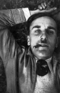 Black and white portrait from a low angle: middle-aged man (Lucien Dubé) lying on his back, looking directly at the camera. Dark hair, receding hairline, thin mustache. Shirt, jacket, and tie. Cigar between his lips. Left arm bent above his head, hand touching his hair.