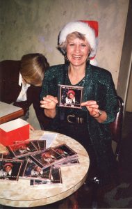 Colour photograph of a blonde haired smiling woman in a dark green sequined dress wearing a red and white Santa Claus hat seated before a round table strewn with compact disc covers holding in front of her a Compact Disc cover bearing her photograph and the title “The Christmas Lady”.