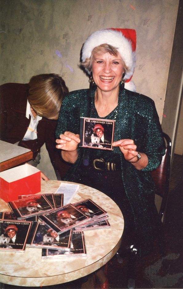 Colour photograph of a blonde haired smiling woman in a dark green sequined dress wearing a red and white Santa Claus hat seated before a round table strewn with compact disc covers holding in front of her a Compact Disc cover bearing her photograph and the title “The Christmas Lady”.
