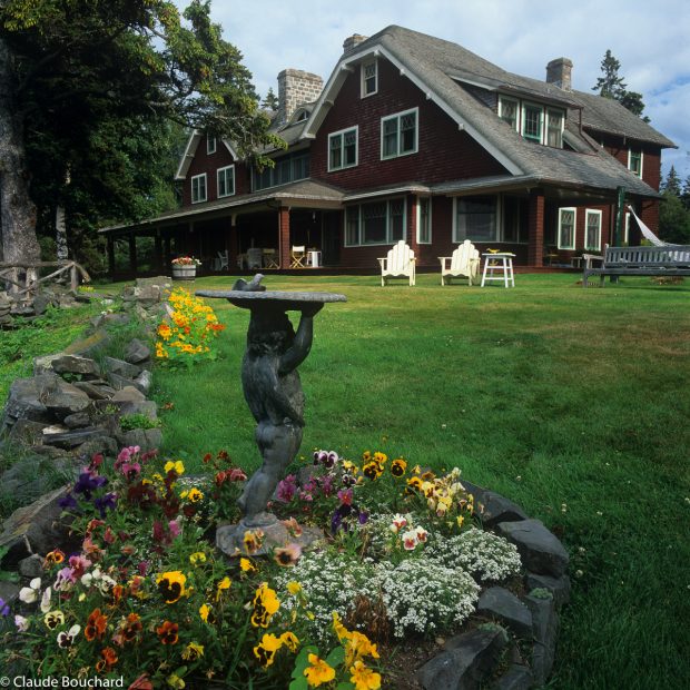 Colour photograph of the façade of a summer home in Métis-sur-Mer. In the foreground, a sculpture of a cherub carrying a birdbath on its head. The sculpture is set in the center of a flowerbed of yellow, orange, white, mauve and pink pansies. The residence is clad in burgundy-coloured cedar shingles. The 2-storey residence has a natural cedar-shingle roof and a covered veranda on the façade. 2 white Ardirondack chairs are placed side by side on the lawn.
