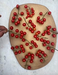 Overhead view of a light wooden oval tray on which numerous clusters of small red cherry tomatoes are arranged, with a hand holding a cluster on the left side.