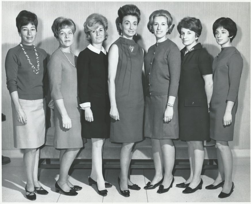 A black-and-white formal photograph of a group of women in 1960s-style attire.
