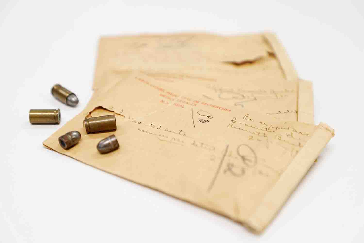 Colour photograph of small caliber rifle cartridges. 2 empty cartridges and 2 rifle bullets and one cartridge still loaded with its bullet. The exhibits are laid flat next to 3 brown 5 x 7 inch envelopes. The short depth of focus makes it impossible to read the writing on the envelopes.