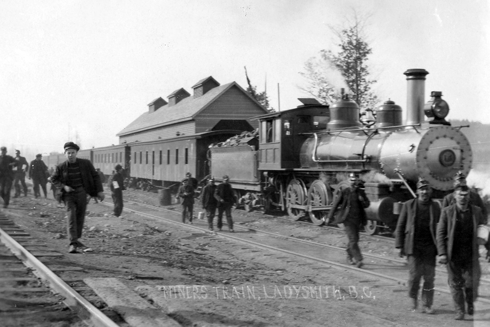 Black and white photograph featuring men walking from a train headed by a steam locomotive.