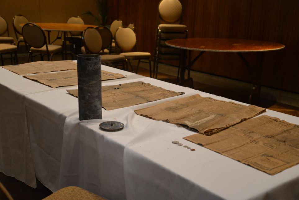 Several old newspapers, a brass cylinder time capsule, and five coins laid out on a table with a white tablecloth.