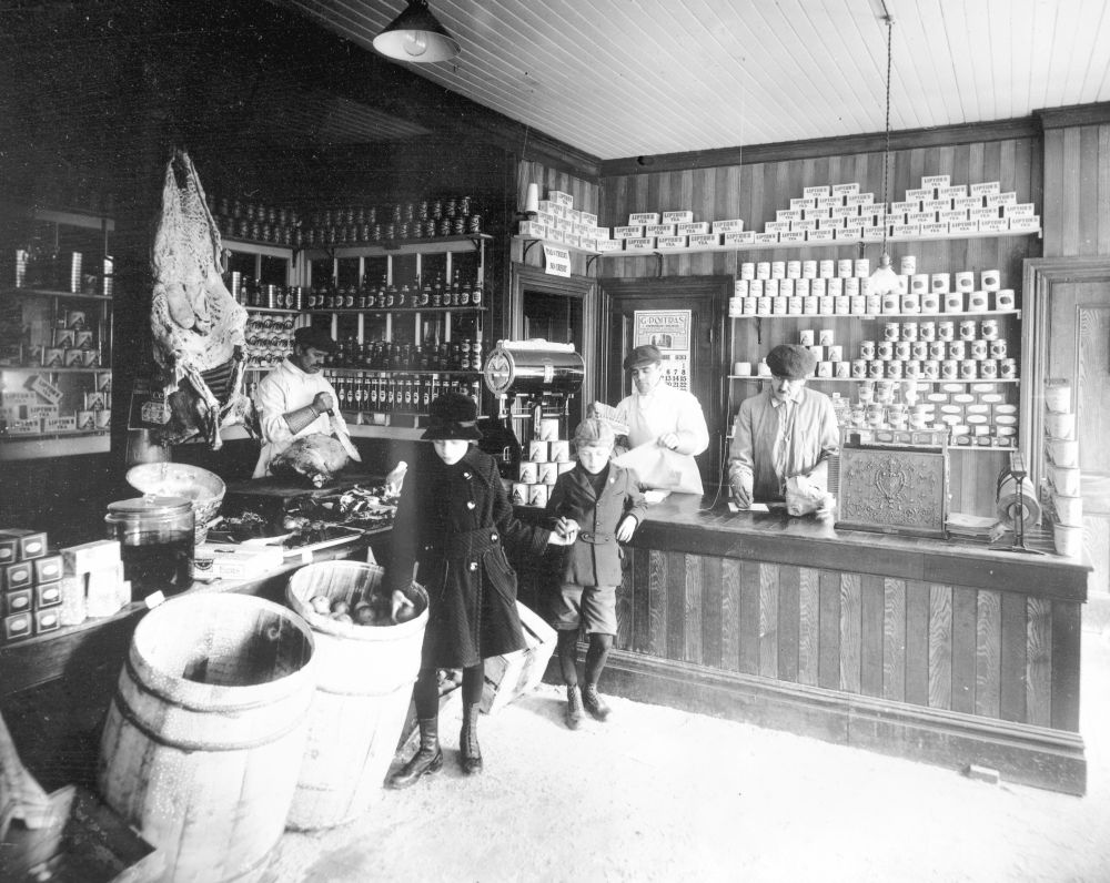 The interior of a 1930s grocery store, two children are taking apples from a barrel, clerks are behind a counter