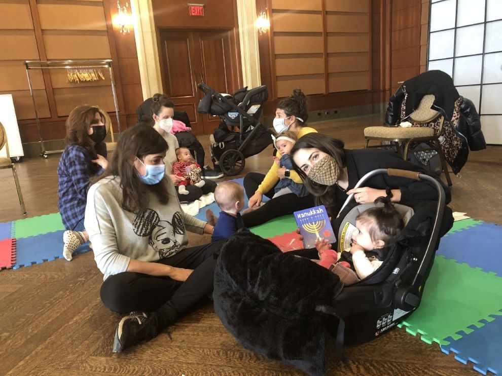 A group of women and children sitting on the floor of a social hall wearing masks during a Shabbat program.
