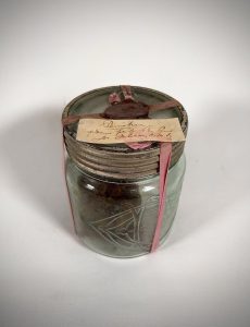 Clear cylindrical glass jar filled with pemmican. Screw-on metal lid. Yellowed paper (handwritten note in brown ink) attached to the lid with a faded pink ribbon and red wax seal.