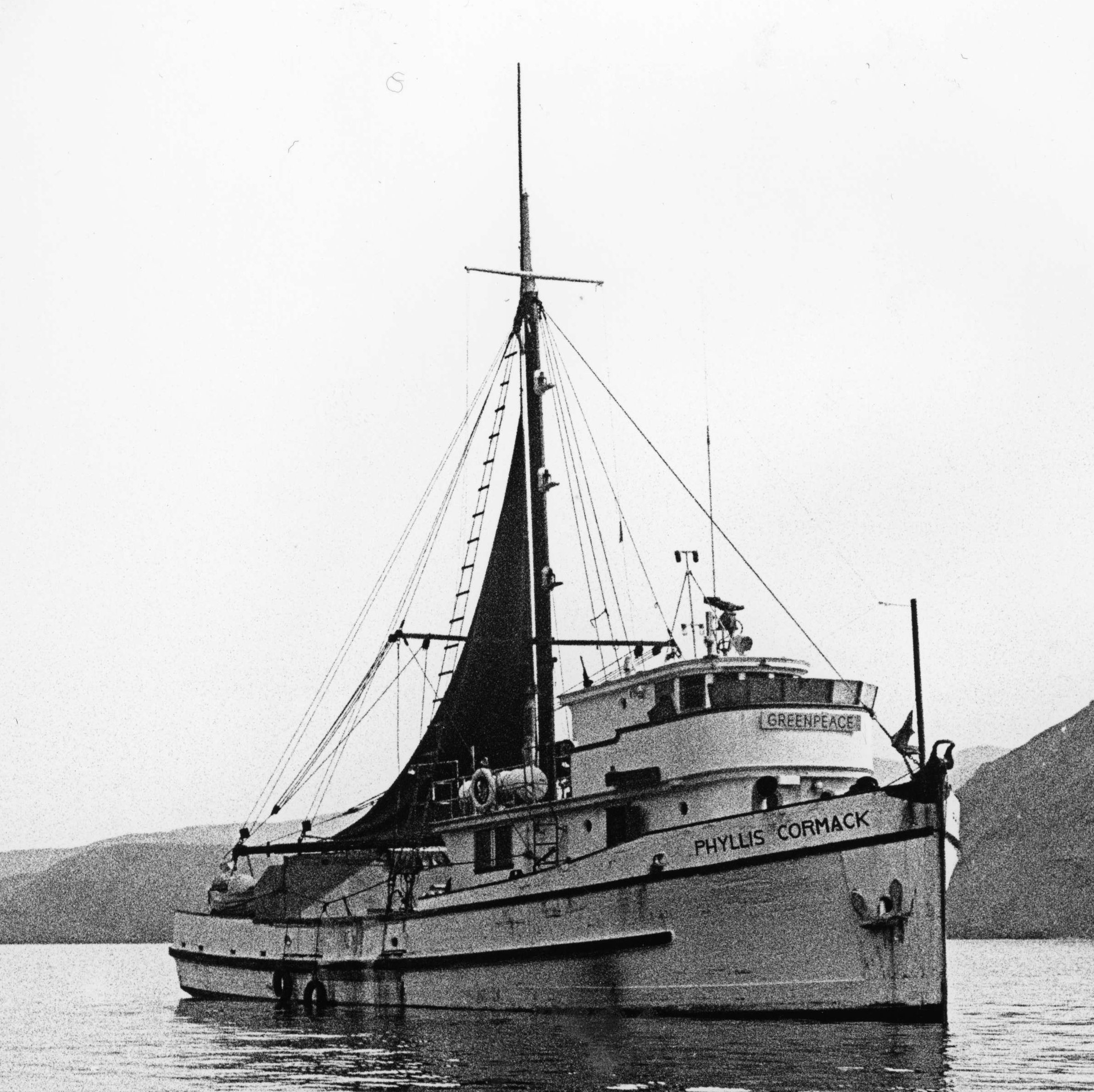 A black and white photograph of a white medium-sized boat sailing on the water with two mountains behind it. On the boat are two names, Phyllis Cormack and Greenpeace, representing the original name and new name.
