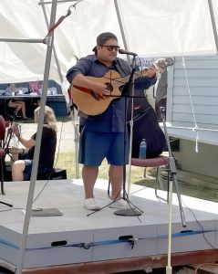 Colour photograph of a Coast Salish musician standing on a stage in front of a microphone playing an acoustic electric guitar and singing.