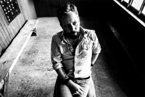 Black and white portrait photograph of a seated bearded man, hair cut above the ears and parted on the left wearing a wrangler style shirt, long sleeves rolled up to the elbows, and denim jeans.