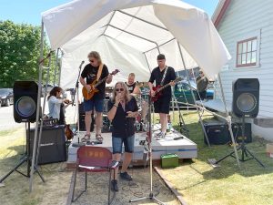 Colour photograph of a five-piece rock band performing on a small outdoor stage shielded from the sun by a white tent.