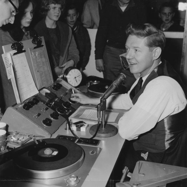Black and white photograph showing a smiling DJ playing records surrounded by a group of admiring teenagerss watch him.