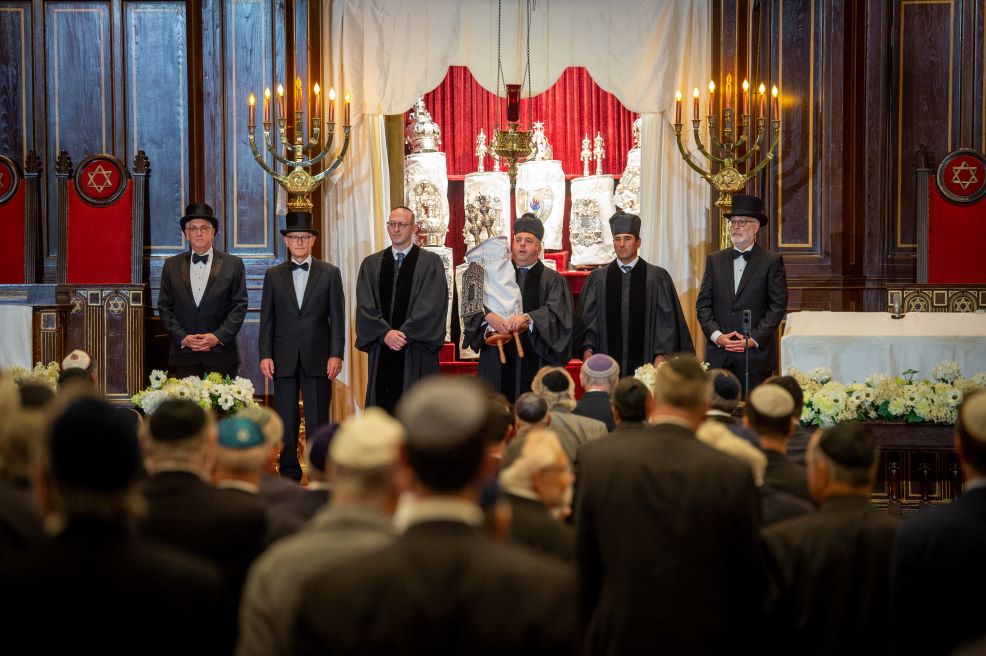 A group of men dressed in formal and ceremonial standing on a synagogue bimah for a rededication ceremony.