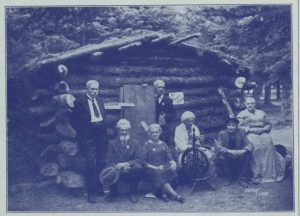 A clipping from a newspaper of a black and white photo of a group of elderly people standing in front of a log cabin. Some are standing and some are seated. One woman is spinning wool.