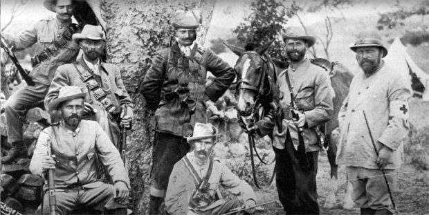 Black and white photograph showing seven men posing with rifles ready for battle. A horse and a tent are in the background.