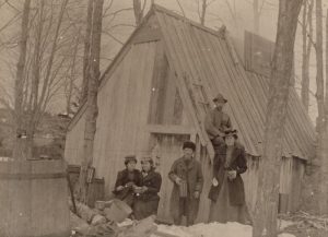 Sepia photograph: family celebration in front of a rustic wood cabin (wooded and snowy environment). Left foreground: two young women sitting. Centre: young man standing. Right: woman standing. Cabin roof: seated man . Far left: large wooden barrel.