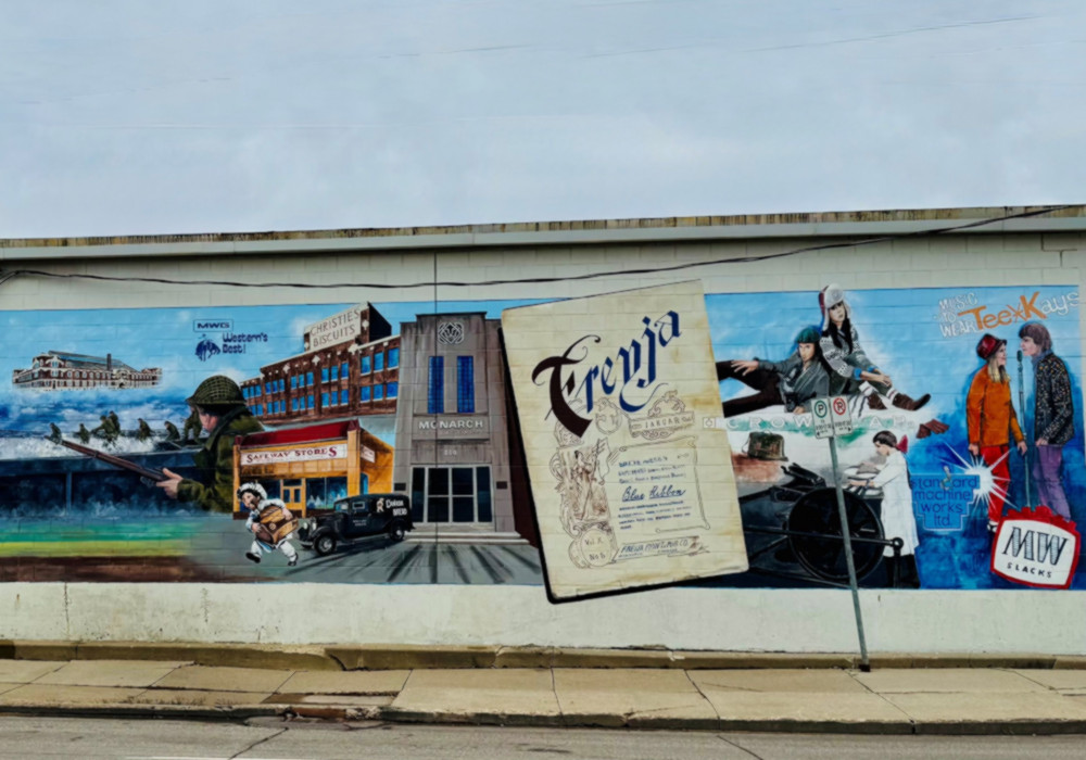 A colourful mural painted on the side of a building depicts scenes from Icelandic-Canadian history. On the left is a red brick factory marked Gimli Fish, beside a river and small figure in a white cap. At the centre stands the Icelandic newspaper building Lögberg-Heimskringla with a vintage car in front. To the right, a large image of the literary magazine Freyja shows its decorative title page, while a motorcyclist and man in old-fashioned clothing appear at the far right.