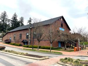 Oblique colour photograph of a two-story rectangular gable-roofed building built into a slight slope, the lower story being below grade at the upper end of the building. Walls are clad with medium brown wooden shakes and feature four rectangular ‘portrait’ windows in the upper story. The roof is dark grey asphalt shingles. At the downslope end a prominent set of steps leads to the upper story entrance.