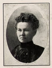 Black-and-white oval-framed portrait of a woman with a serious expression. She has curly hair styled up and wears a high-collared dark dress with pleated detailing. The photograph has a formal, studio-style appearance.